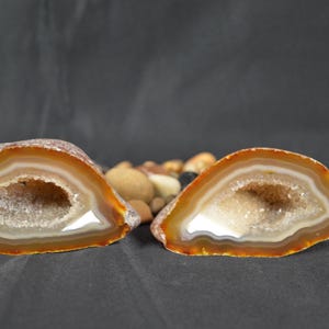 May include: Two halves of a geode, displaying a natural, layered structure. The outer edges are a warm orange-brown, fading into white and a sparkling crystal centre. The cut reveals a hollow interior.