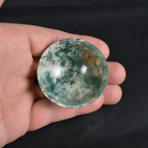 Puede incluir: Un cuenco redondo de piedra pulida con un patrón moteado de tonos verdes, blancos y anaranjados. El cuenco se sostiene en una mano, mostrando su superficie lisa y sus colores naturales. El diámetro del cuenco es de aproximadamente 5 cm.