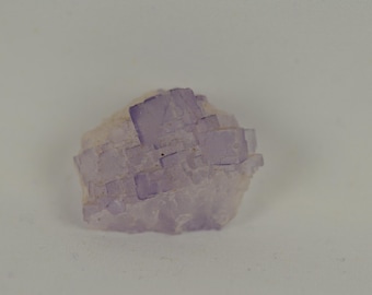 以下が含まれることがあります： 天然の蛍石結晶標本。結晶は、淡い紫色の色合いと粗くテクスチャ加工された表面を持つ、層状の構造を特徴としています。結晶は無地の白い背景に配置されています。