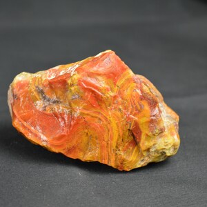 May include: A natural, uncut stone with a vibrant colour palette. The rock features layers of red, orange, and yellow hues, creating a swirling pattern. The stone's surface is rough and textured, showcasing its natural form. The background is a solid black.