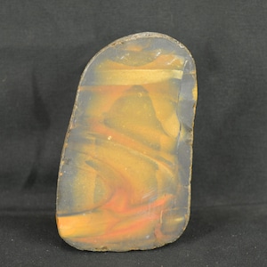 以下が含まれることがあります： 黄色、オレンジ、グレーの色合いが渦巻く模様の、磨かれた長方形の石。石は滑らかで光沢のある表面を持ち、自然で不規則な形をしています。色が混ざり合い、ユニークで芸術的な外観を作り出しています。