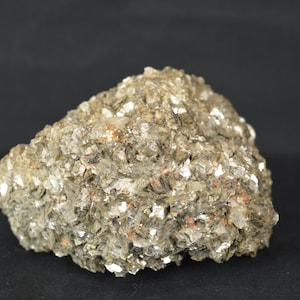 May include: A textured rock formation, predominantly grey and brown, with reflective white crystalline flakes. The surface is uneven and layered. The rock is set against a dark background. The overall appearance suggests a mineral specimen.