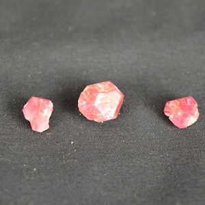 May include: Three raw, faceted, pink gemstones are displayed against a dark gray background. The gemstones vary in size, with the largest in the center and the smallest on the left. The gems have a rough, natural texture.