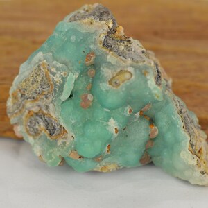 May include: A close-up of a raw, uncut mineral specimen. The stone features a vibrant turquoise-green hue with a textured, bumpy surface. Patches of tan and gray are visible, adding contrast. The mineral sits on a wooden surface.