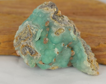 Puede incluir: Primer plano de un espécimen mineral en bruto sin cortar. La piedra presenta un vibrante tono verde turquesa con una superficie texturizada y rugosa. Se ven parches de color beige y gris, añadiendo contraste. El mineral está sobre una superficie de madera.
