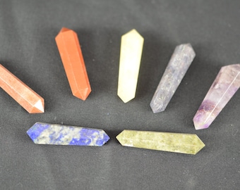 May include: Assortment of seven gemstone wands in various colors, including red, orange, yellow, blue, green, gray, and purple. The wands are hexagonal and have pointed ends. The background is a dark gray.