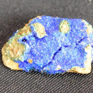 May include: A vibrant blue azurite mineral specimen with patches of green and tan hues. The stone has a rough, textured surface and is displayed against a dark background. This natural stone is a beautiful example of a mineral.