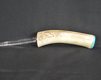 May include: A handcrafted knife with a curved antler handle and a clear, pointed blade. The handle features a light brown and cream color with a turquoise accent at the end. The knife is set against a black background.