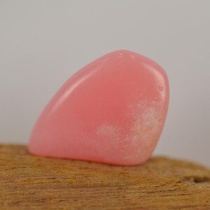 Peut inclure: Une pierre précieuse rose polie et arrondie. La pierre a un aspect doux et translucide avec une zone plus claire et opaque sur un côté. Elle repose sur un morceau de bois brun texturé.