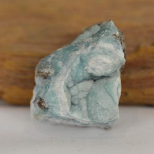 May include: A close-up of a light blue mineral specimen with a textured, bubbly surface. The stone has areas of white and brown, and is set against a blurred wooden background.