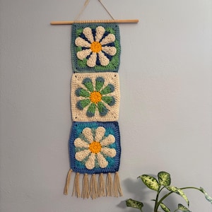 May include: A handmade crochet wall hanging featuring three square panels with daisy designs. Each panel has a different color scheme, with blue, green, and cream backgrounds. The daisies have white petals and yellow centers. The wall hanging is suspended from a wooden dowel.