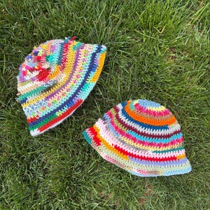 May include: Two colorful crocheted bucket hats with a variety of stripes in shades of blue, green, red, yellow, orange, and pink. The hats are lying on a bed of green grass.