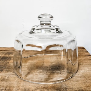 Può includere: Una campana di vetro trasparente con forma ottagonale arrotondata e un piccolo pomello sulla parte superiore. La campana è progettata per coprire e proteggere gli alimenti. Si trova su una superficie di legno.