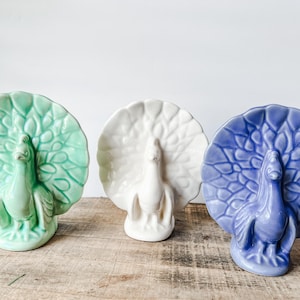 May include: Three ceramic peacock figurines in shades of green, white, and blue. Each peacock has a detailed tail and stands on a wooden surface. The figurines are decorative and add a touch of elegance.