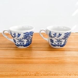 May include: Two white teacups with blue Willow pattern designs. Each cup has a curved handle and a detailed landscape scene with birds, trees, and a bridge. The cups are placed on a light brown wooden surface.