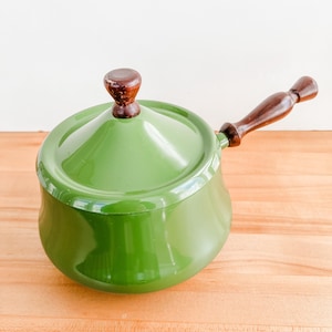 May include: A green enamel saucepan with a lid and a wooden handle. The saucepan is a vibrant green color and has a dark brown wooden handle and lid knob. The pot is sitting on a wooden surface.