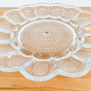 May include: Clear glass serving platter with a scalloped edge and a central circular area. The platter features a textured pattern of small circles in the center and a floral design around the edge. The platter is on a wooden surface.