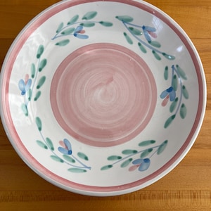 May include: White ceramic plate with a pink center and rim. The plate features a floral design with blue flowers and green leaves. The plate is round and has a slightly raised edge.