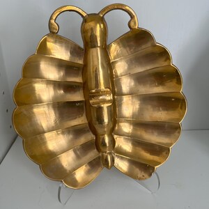 May include: A polished gold-colored metal dish shaped like a butterfly. The dish features a central body section with two symmetrical, scalloped wings on either side. The surface has a smooth, reflective finish with visible metallic texture.