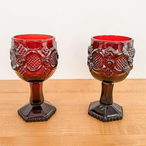 Può includere: Due calici vintage rosso rubino con disegni in rilievo ornati. I bicchieri hanno un gambo corto e una base esagonale. I calici sono in vetro e sono esposti su una superficie di legno chiaro.