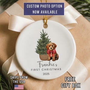 May include: A white ceramic Christmas ornament featuring a puppy with a red bow and a Christmas tree. The text reads "Frankie's First Christmas 2025". The ornament has a gold string for hanging. The text "CUSTOM PHOTO OPTION NOW AVAILABLE" is at the top.