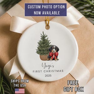 May include: A white ceramic Christmas ornament with a watercolor illustration of a puppy wearing a red bow, next to a Christmas tree. The text reads "Yogi's First Christmas 2025". The ornament has a gold string for hanging. The text "CUSTOM PHOTO OPTION NOW AVAILABLE" is at the top.