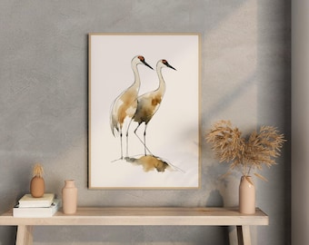 May include: Watercolor painting of two cranes with brown and beige plumage, standing side-by-side. The birds have long, thin legs and red markings on their heads. The artwork is framed in a light wood frame.