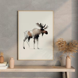 Puede incluir: Pintura a la acuarela de un alce enmarcada. La obra presenta un estilo minimalista con trazos de tinta negros y marrones. El alce está representado de pie sobre un fondo crema. El marco es de madera.