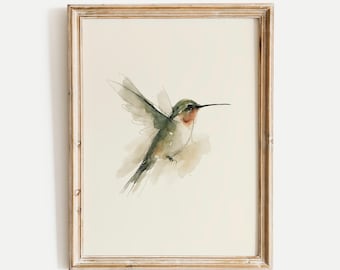 May include: A framed watercolor painting of a hummingbird in flight, featuring soft green, grey, and reddish-orange tones on a cream-colored background. The artwork is enclosed in a light-toned wooden frame with a visible grain and a simple profile.