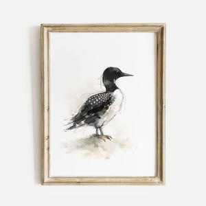 May include: Watercolor print of a loon bird in a wooden frame. The bird is depicted in black and white with a speckled pattern on its back and a dark head. The artwork has a minimalist style and is set against a white background.