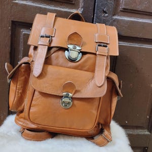 Puede incluir: Una mochila de cuero marrón con dos bolsillos grandes en la parte delantera y dos correas ajustables. La mochila tiene dos hebillas metálicas y dos cierres metálicos.