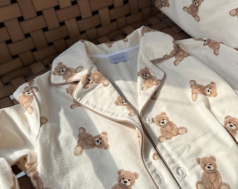 May include: Cream-colored pajama set with a repeating pattern of brown teddy bears. The set includes a button-down shirt with a notched collar and a matching pillow. The word "BELLA" is printed on the shirt's label.