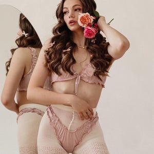 Puede incluir: Una mujer con un conjunto de lencer&iacute;a rosa y medias de encaje a juego. Lleva flores en el pelo y sostiene un ramo de rosas rosas.