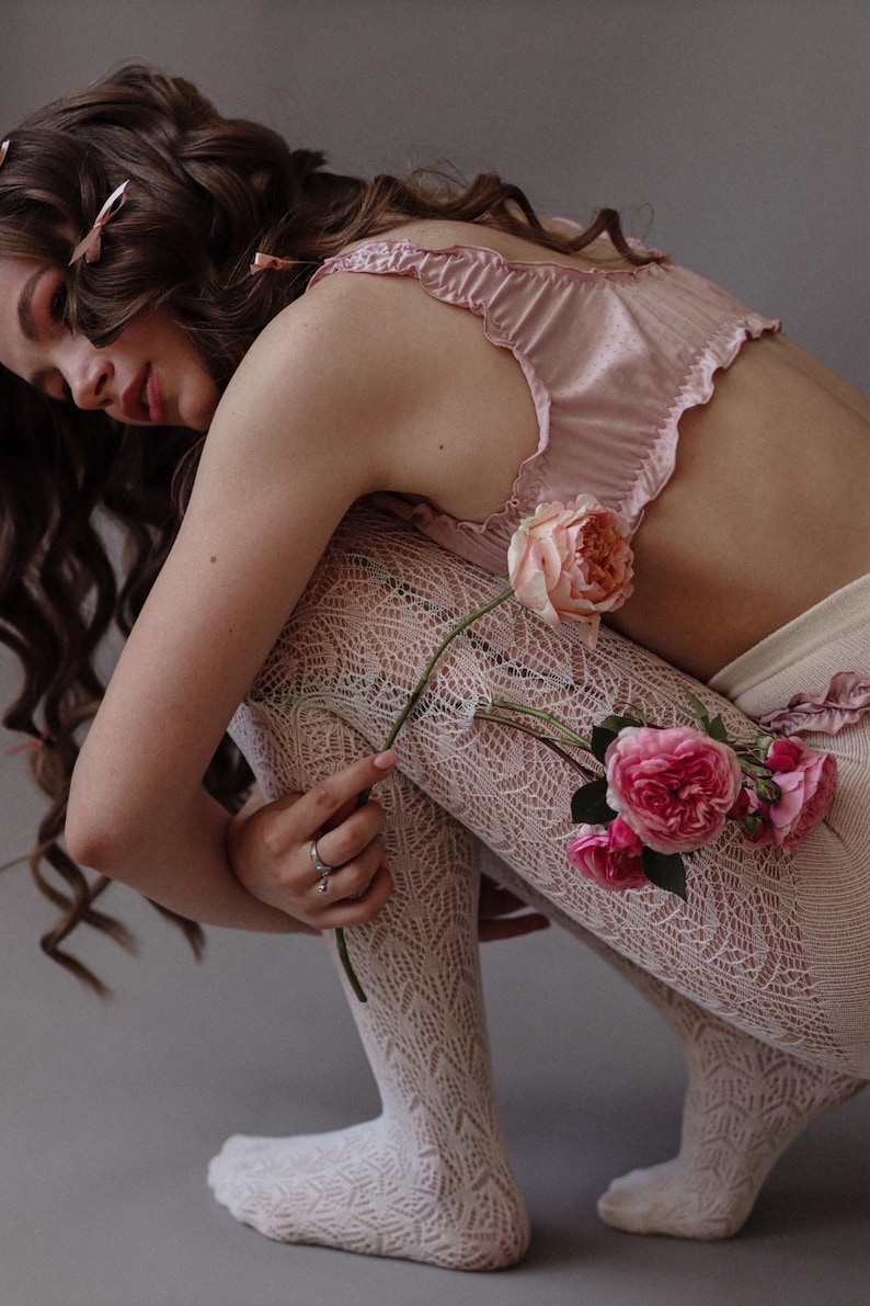 Puede incluir: Una mujer con un top de sat&eacute;n rosa y medias de encaje blanco posa con un ramo de rosas rosas.