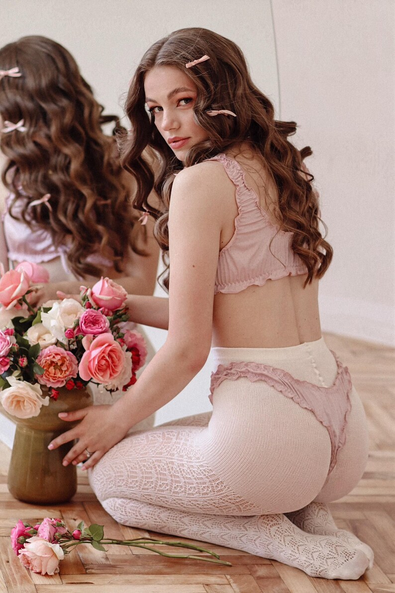 Puede incluir: Una mujer con un conjunto de lencer&iacute;a rosa con ribete de encaje blanco y medias de encaje blanco. Est&aacute; sentada en un suelo de madera con un ramo de rosas rosas delante de ella.
