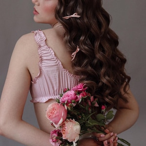 Puede incluir: Una mujer con el pelo largo, casta&ntilde;o y ondulado lleva una blusa de sat&eacute;n rosa con ribete de volantes y sostiene un ramo de rosas rosas.