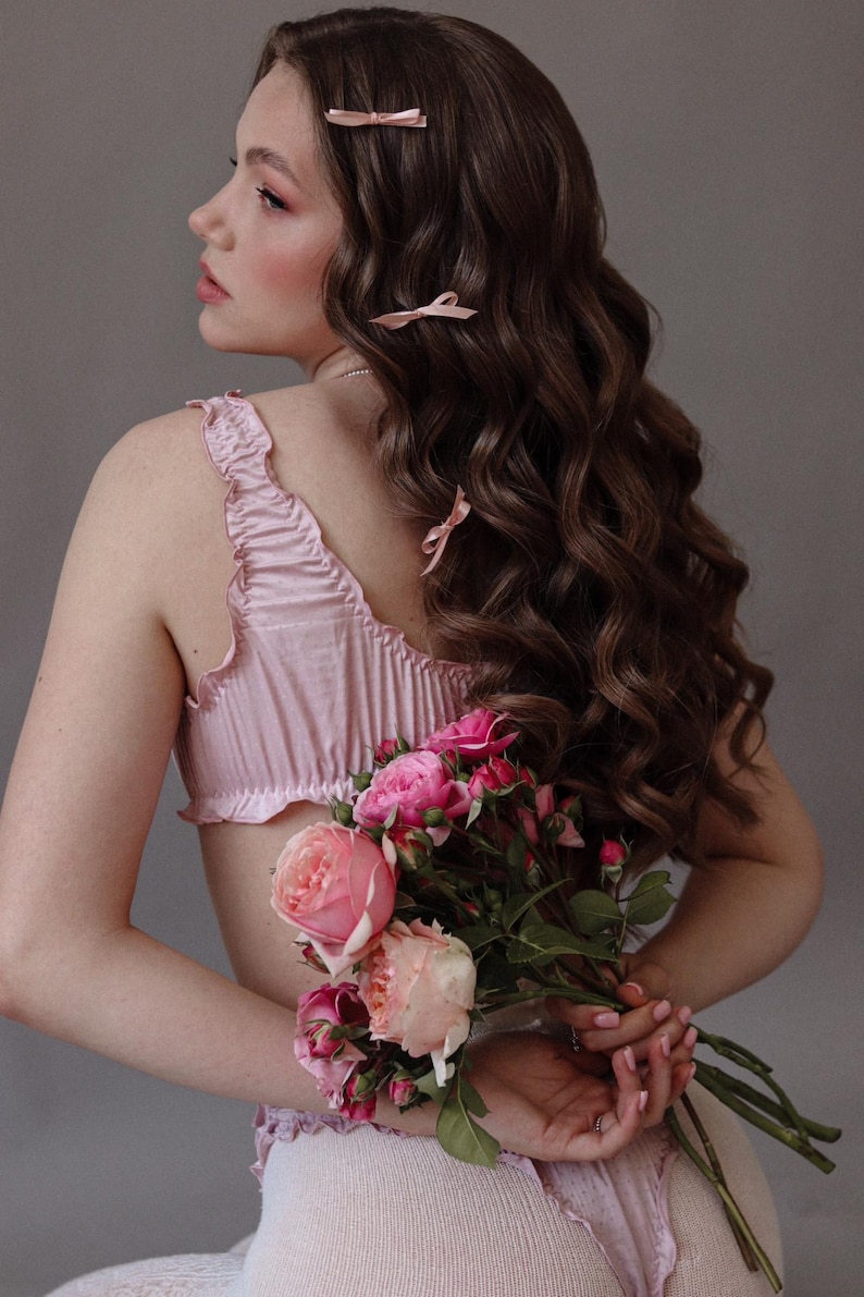 Puede incluir: Una mujer con el pelo largo, casta&ntilde;o y ondulado lleva una blusa de sat&eacute;n rosa con ribete de volantes y sostiene un ramo de rosas rosas.