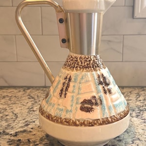 May include: A vintage ceramic pitcher with a cream-colored base and a conical body decorated with brown, blue, and pink patterns. It features a silver-colored metal top and handle. The pitcher is likely for serving beverages.