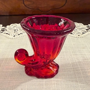 May include: A ruby red glass vase with a unique, curved design. The vase has a flared top, a rounded, curled accent, and a textured base. The glass is transparent, allowing light to pass through, and sits on a neutral-colored surface.
