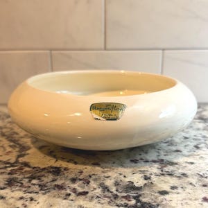 May include: A low-profile, cream-colored ceramic bowl with a smooth, rounded rim. A gold-toned, rectangular sticker is affixed to the front, featuring the text Haeger Flora, with additional smaller text below indicating it was made in the U.S.A.