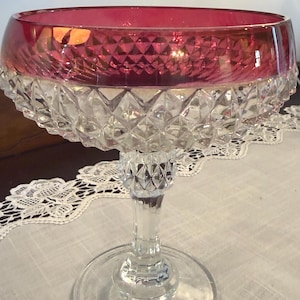 May include: A vintage champagne coupe with a ruby red rim and a clear, diamond-cut bowl. The stem and base are also clear glass. The glass sits on a white lace tablecloth.