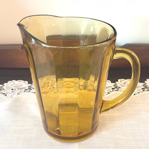 May include: Amber-colored glass pitcher with a handle and a pouring spout. The pitcher has a ribbed design and sits on a white lace tablecloth. The pitcher is empty and appears to be vintage.