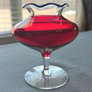May include: A clear glass vase with a ruffled rim, filled with a vibrant red liquid. The vase has a bulbous body and a slender stem, resting on a circular base. The background is blurred, suggesting an indoor setting.