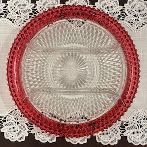 May include: A clear glass divided serving dish with a ruby red textured rim. The dish has three sections and a textured center. It sits on a white lace-trimmed cloth.