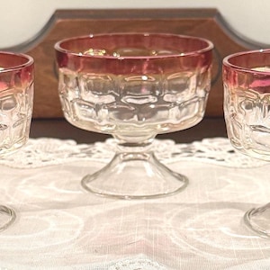 May include: Three vintage clear glass goblets with a red-orange rim and a textured, honeycomb-like pattern. The goblets have a short stem and a wide, shallow bowl. They are arranged on a white lace tablecloth.
