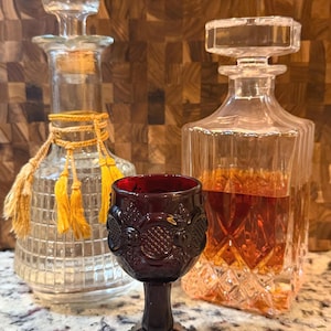 May include: A ruby red glass with ornate detailing stands before two glass decanters. One decanter is filled with amber liquid. The other is empty and has a cork stopper. The decanters and glass are on a speckled surface.