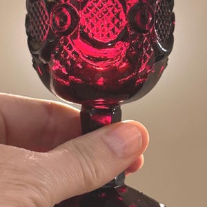 May include: A ruby red, vintage-style glass goblet. The goblet features an ornate, embossed design around the bowl and a detailed base. The stem is short and the glass has a glossy finish. The goblet is held in a hand.