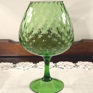 May include: A green glass goblet with a textured surface and a wide, rounded bowl. The goblet has a stem and a circular base. The rim of the bowl has a scalloped edge. The glass has a slight iridescence.