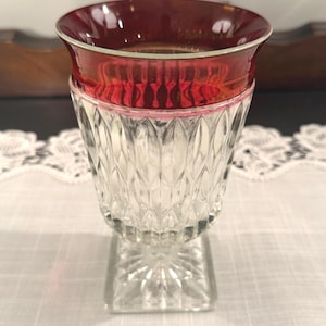 May include: A clear glass goblet with a ruby red rim and a decorative, textured pattern. The goblet sits on a square base. The rim has a thin, white border. The glass has a vintage aesthetic.
