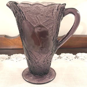 May include: A vintage purple glass pitcher with a pear and leaf design. The pitcher has a handle and a scalloped rim. The base is round and the pitcher is made of textured glass. The pitcher is likely used for serving beverages.